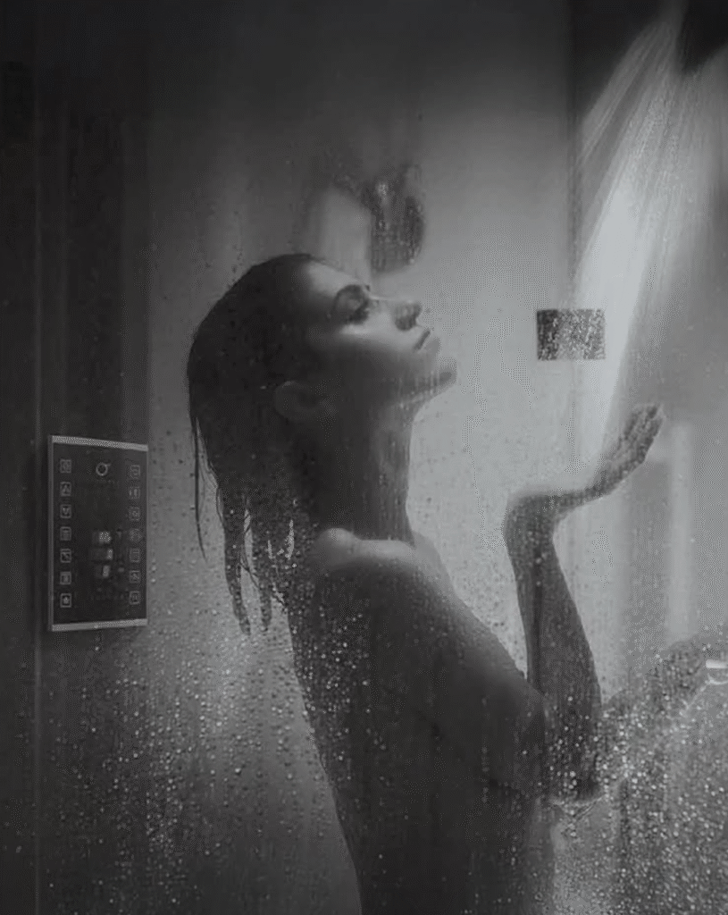 Woman enjoying a relaxing rainfall shower inside a modern glass shower enclosure, water droplets surrounding her while a digital shower control panel is visible on the wall, illustrating a premium spa-like bathroom experience.