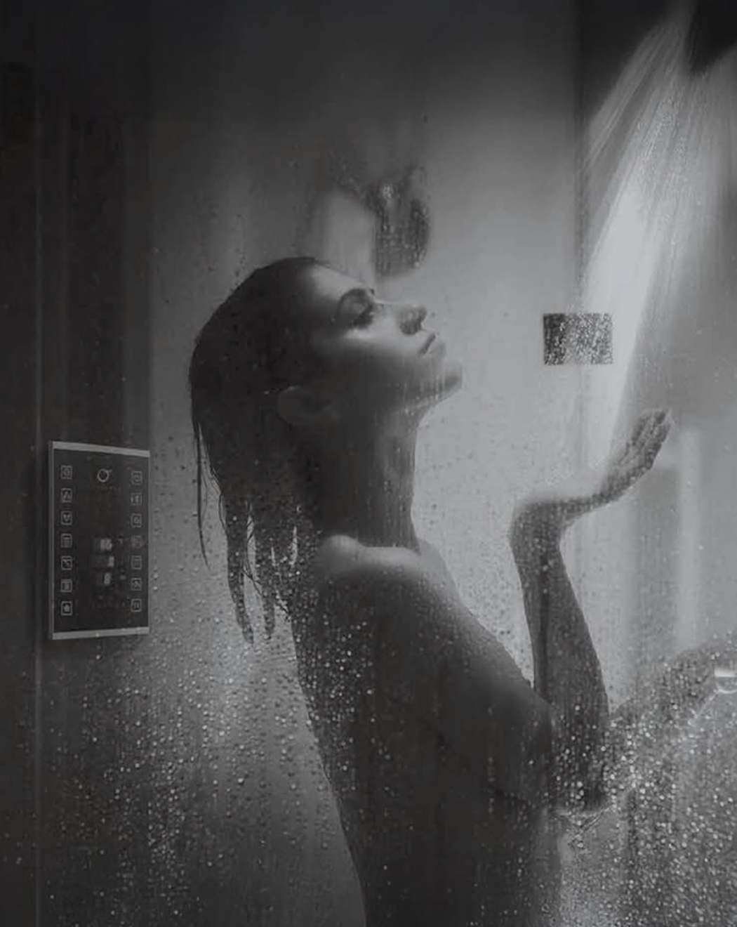 Woman enjoying a relaxing rainfall shower inside a modern glass shower enclosure, water droplets surrounding her while a digital shower control panel is visible on the wall, illustrating a premium spa-like bathroom experience.